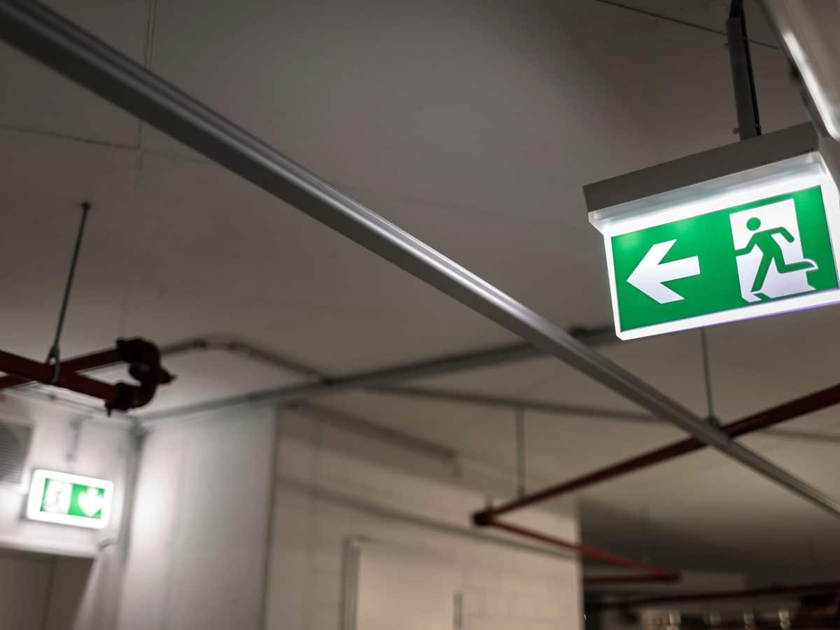Beleuchtetes Rettungswegzeichen in einem Gebäude, im Hintergrund weitere Fluchtwegkennzeichnung und Brandschutztechnik